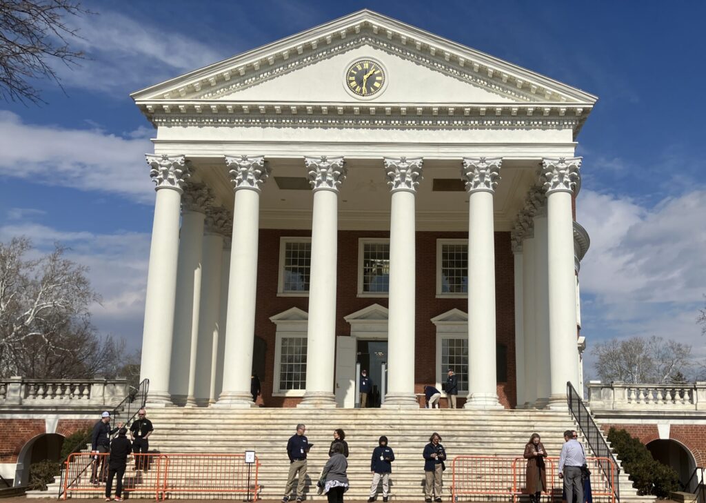 A large brick building with a triangular roof, white columns, and several stairs, with several people waiting behind an orange fence in front of the stairs. 