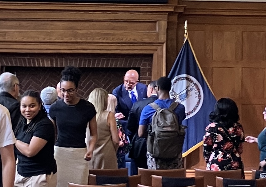 A man in a suit greets a group of people while exiting a room. He is standing in front of an empty fireplace and a blue flag. 