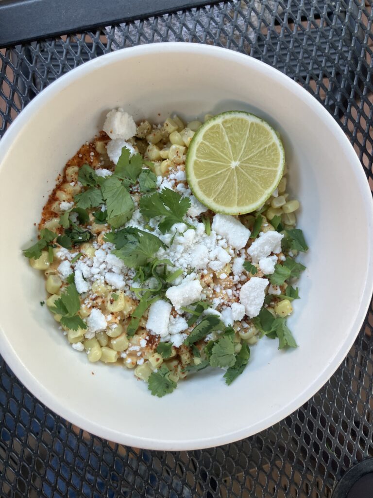 A white bowl of yellow corn, with green herbs, half a lime, spices, and feta crumbles on top.