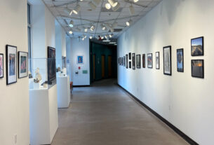 The walls of a hallway are decorated on both sides with PVCC student artwork. On the left side two glass cases containing ceramic works can be seen.