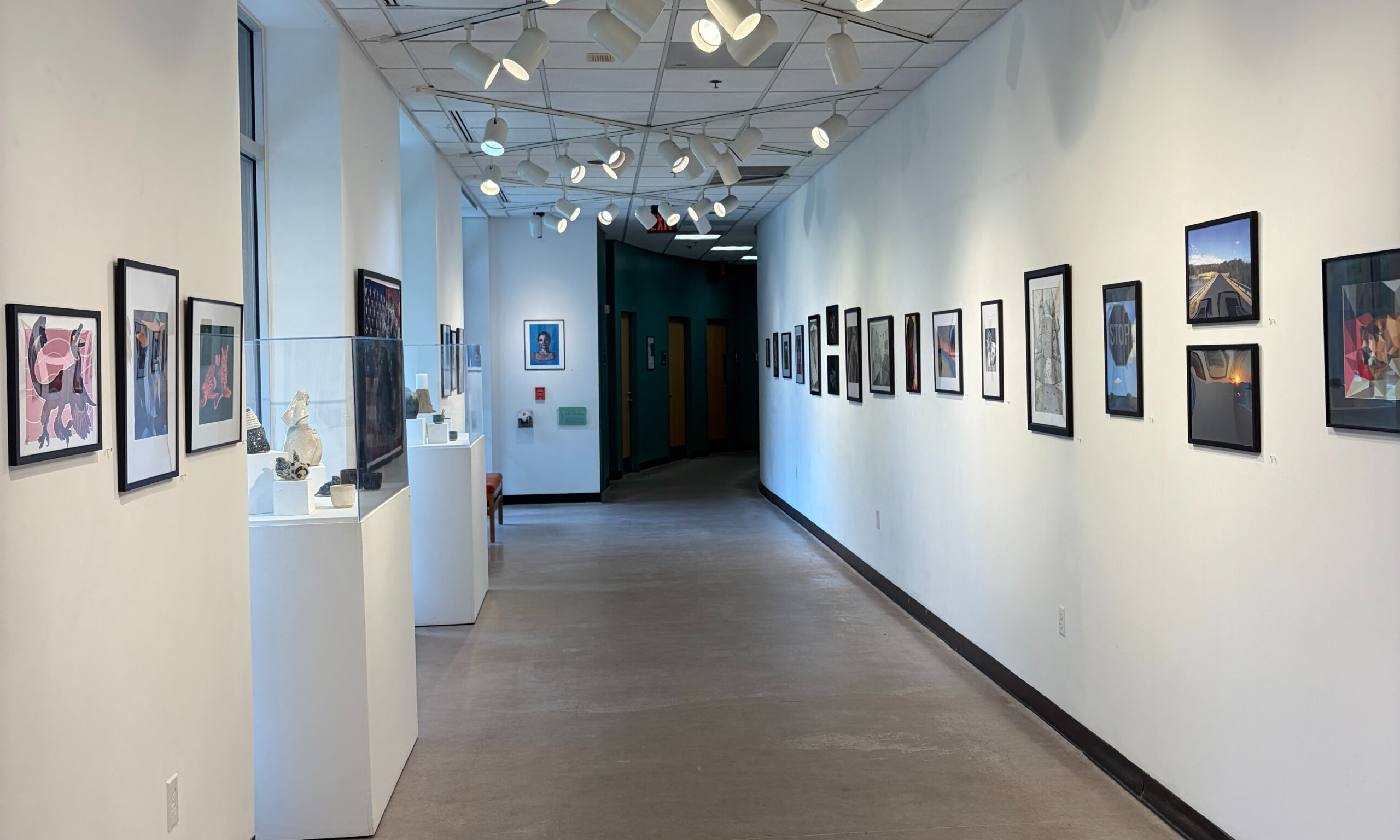 The walls of a hallway are decorated on both sides with PVCC student artwork. On the left side two glass cases containing ceramic works can be seen.