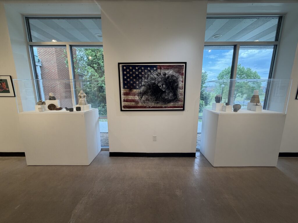A framed mixed-media art piece hangs on the wall. It depicts a distraught-looking head and shoulders of the Statue of Liberty, done in charcoal, on a worn American flag. On both sides of the piece are pedestals with glass cases displaying ceramic works. The pedestals are in front of floor-to-ceiling windows. 