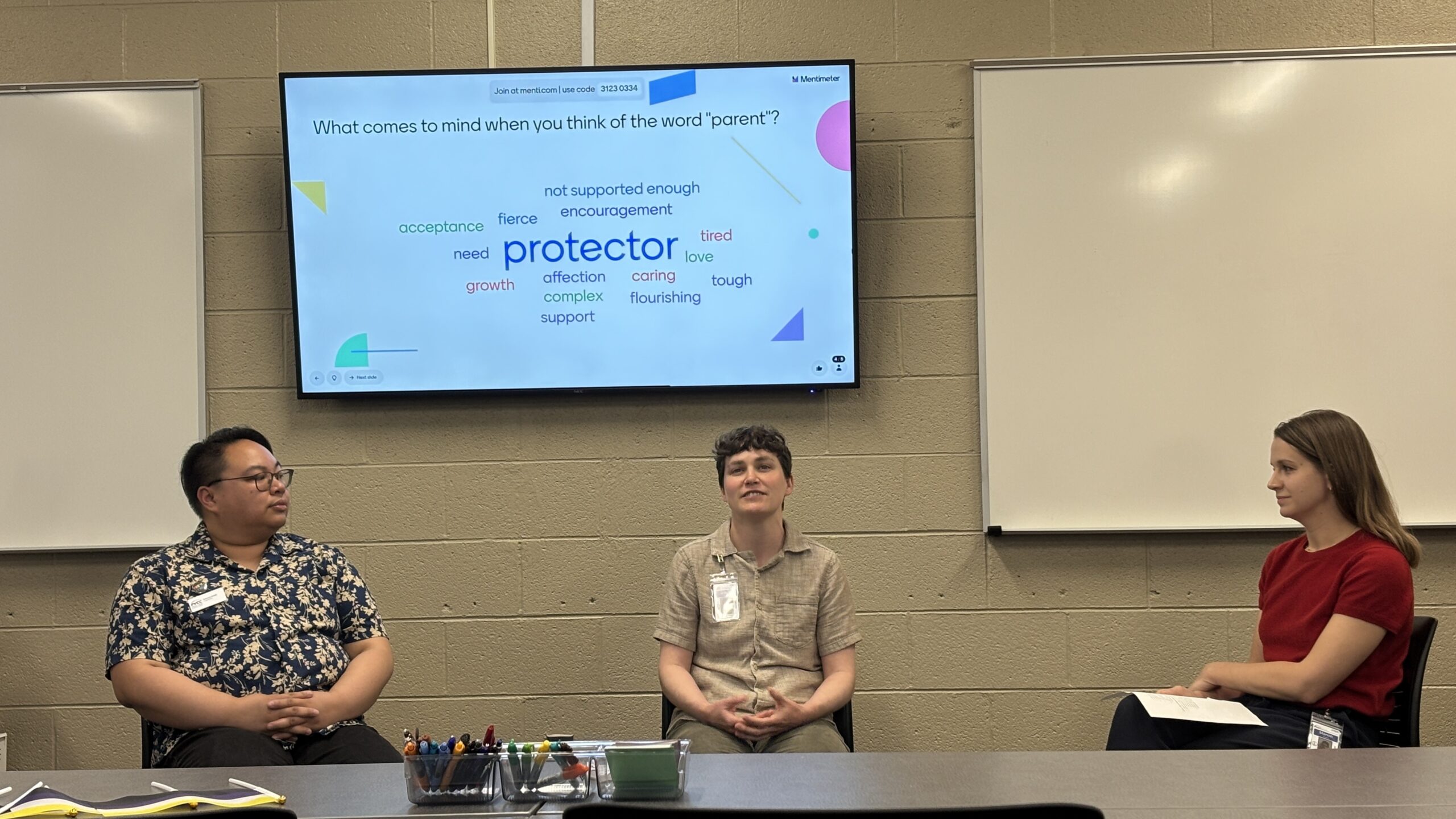 Allyson Umali, Suz Bayker, and Manon Ehrlich sitting behind a table at the panel. Behind them is a TV with a Mentimeter prompt reading "What comes to mind when you think of the word 'parent'?" The largest word on the screen is "protector."