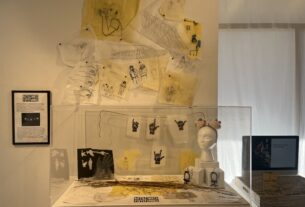 A glass box display containing sheets of paper with hand signs on them, a bust, nettles, and cut outs of paper puppets. More paper characters and drawings hand above.