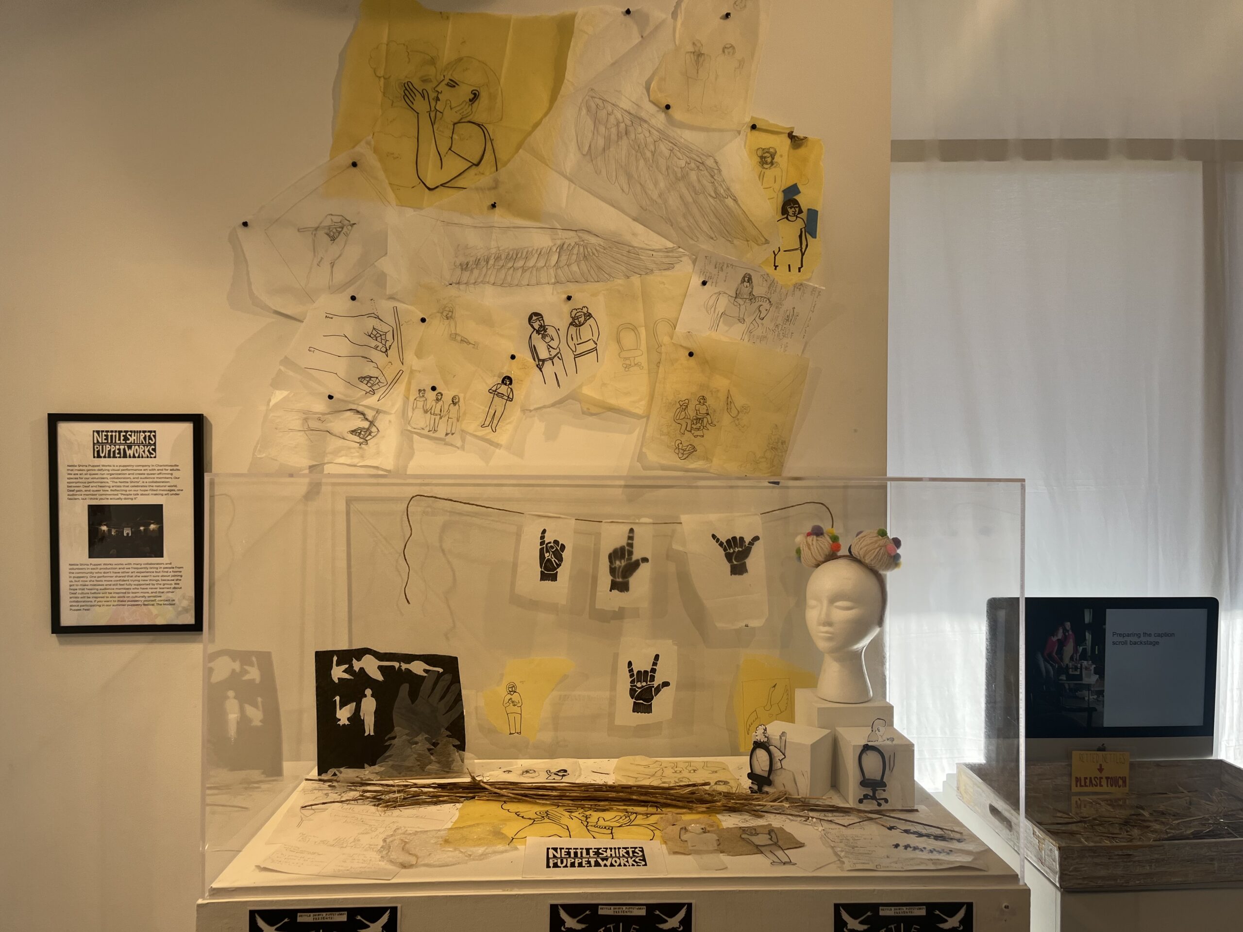 A glass box display containing sheets of paper with hand signs on them, a bust, nettles, and cut outs of paper puppets. More paper characters and drawings hand above.