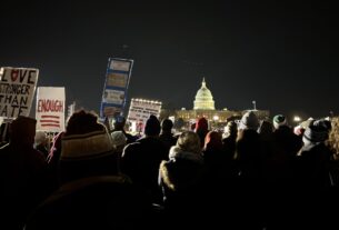 A gathering of attendees pictured from behind a few dozen yards from the stage at night. The Capitol Building sits illuminated in the background. Some attendees are holding signs reading, "LOVE STRONGER THAN HATE," "ENOUGH," "HANDS OFF THE CONSTITUTION," "STAND WITH UKRAINE," "PEOPLE'S STATE OF THE UNION. PEDOPHILE PROTECTORS ARE IN THE WHITE HOUSE!!", other cut-off messages, and images of the Constitution and an upside-down American flag.