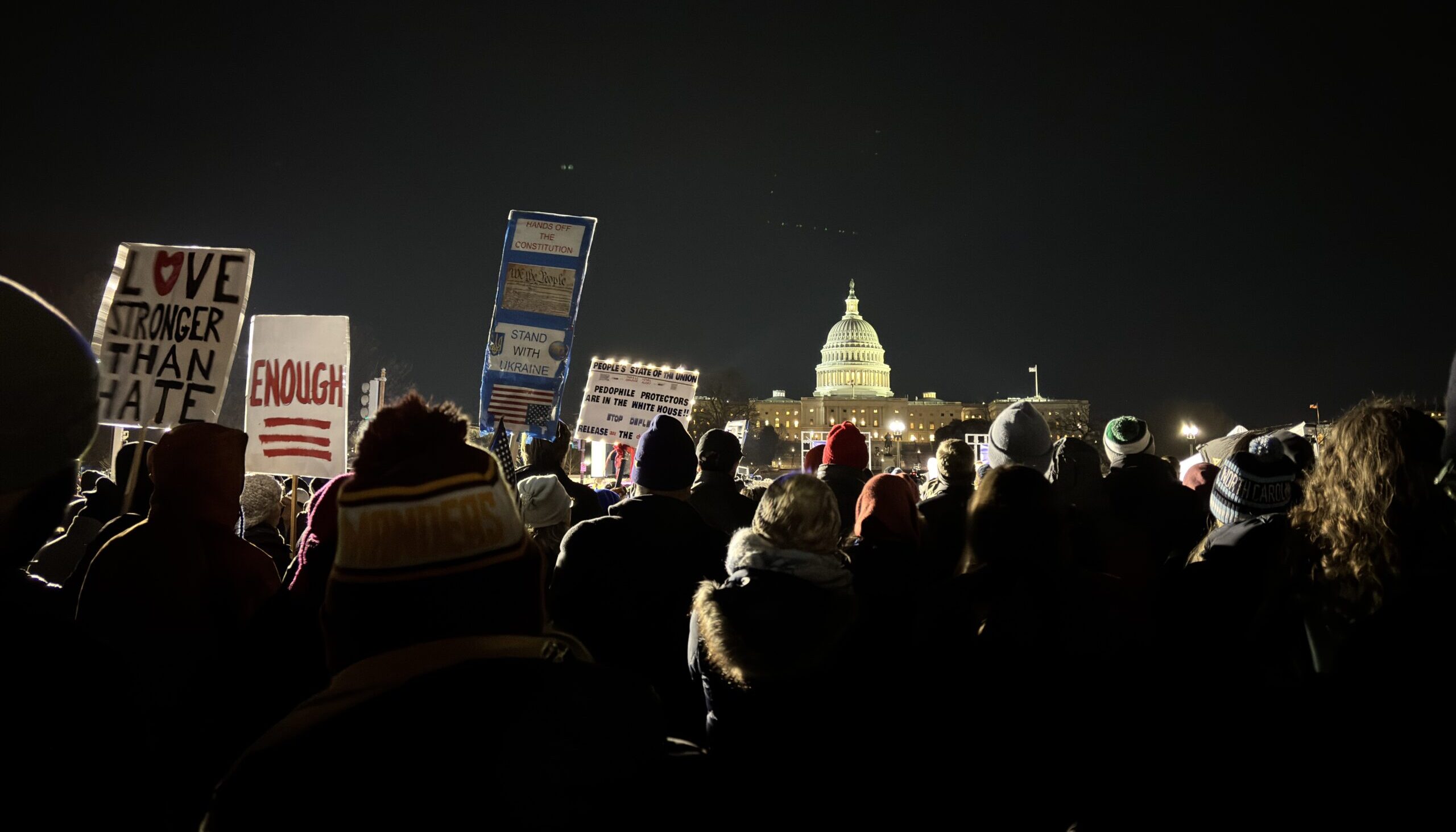 A gathering of attendees pictured from behind a few dozen yards from the stage at night. The Capitol Building sits illuminated in the background. Some attendees are holding signs reading, "LOVE STRONGER THAN HATE," "ENOUGH," "HANDS OFF THE CONSTITUTION," "STAND WITH UKRAINE," "PEOPLE'S STATE OF THE UNION. PEDOPHILE PROTECTORS ARE IN THE WHITE HOUSE!!", other cut-off messages, and images of the Constitution and an upside-down American flag.
