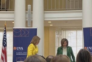 Susan Page, in yellow, and Nancy Pelosi, in green, standing in between two blue banners for the Karsh Institute, next to an American Flag, with columns and windows in the background.