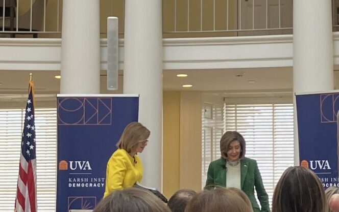 Susan Page, in yellow, and Nancy Pelosi, in green, standing in between two blue banners for the Karsh Institute, next to an American Flag, with columns and windows in the background.