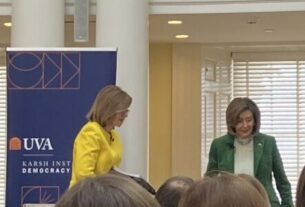 Susan Page, in yellow, and Nancy Pelosi, in green, standing in between two blue banners for the Karsh Institute, next to an American Flag, with columns and windows in the background.