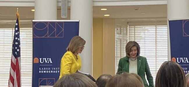 Susan Page, in yellow, and Nancy Pelosi, in green, standing in between two blue banners for the Karsh Institute, next to an American Flag, with columns and windows in the background.