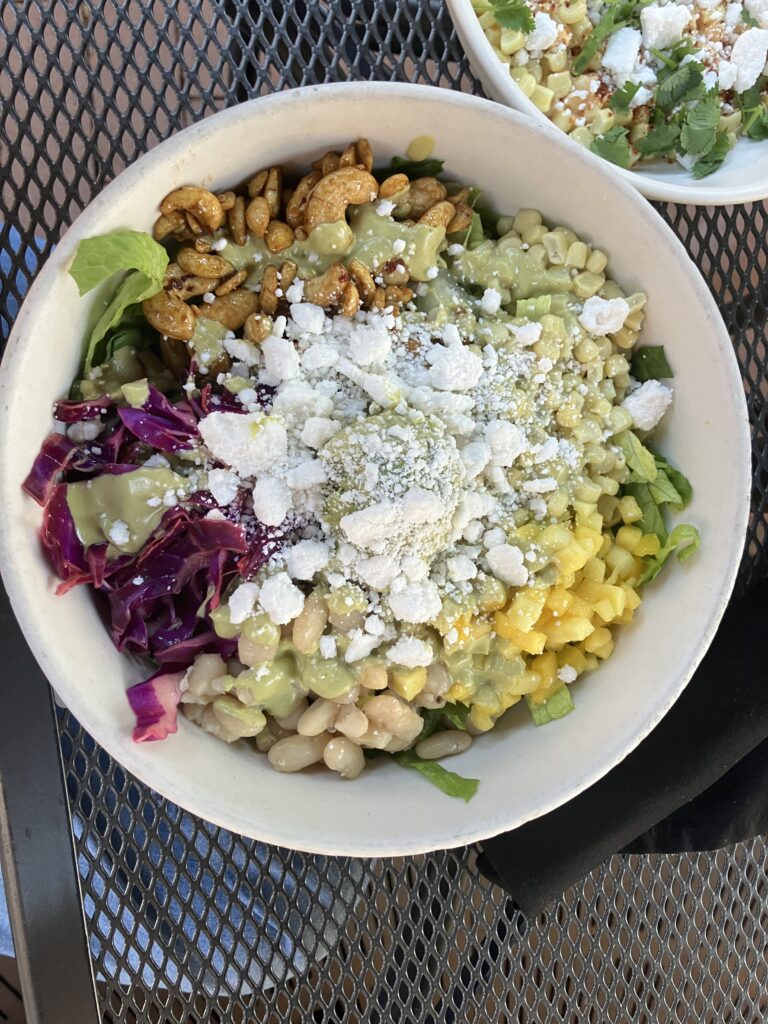 A white bowl with green lettuce, cashews, purple cabbage, white beans, corn, mango, avocado, and crumbled vegan feta. 