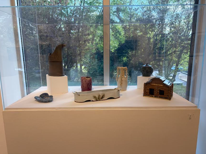 A glass case contains a collection of ceramic works: a bronze colored pitcher, a red-and-white-spotted mug, a white vase with chains connected to the rim, a dark green garlic jar, a small log cabin, an oblong bread tray with a leaf decoration in the center, and a small spotted creature. 