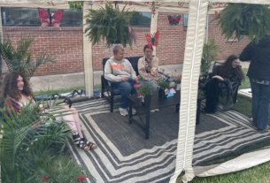 Four students sit on patio furniture in the butterfly tent surrounded by flowers, butterfly cutouts, and hanging plants.