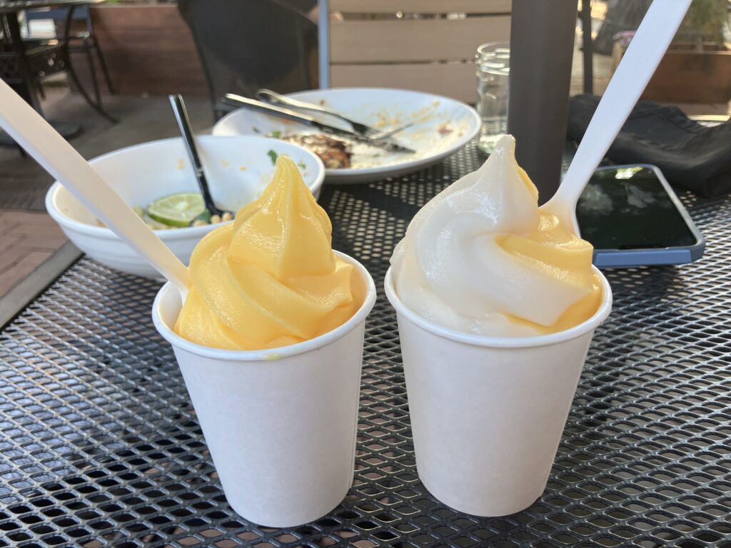 Two white cups, one of yellow ice cream and the other with yellow and white swirl ice cream and both with white spoons, on a table with empty dishes in the background. 