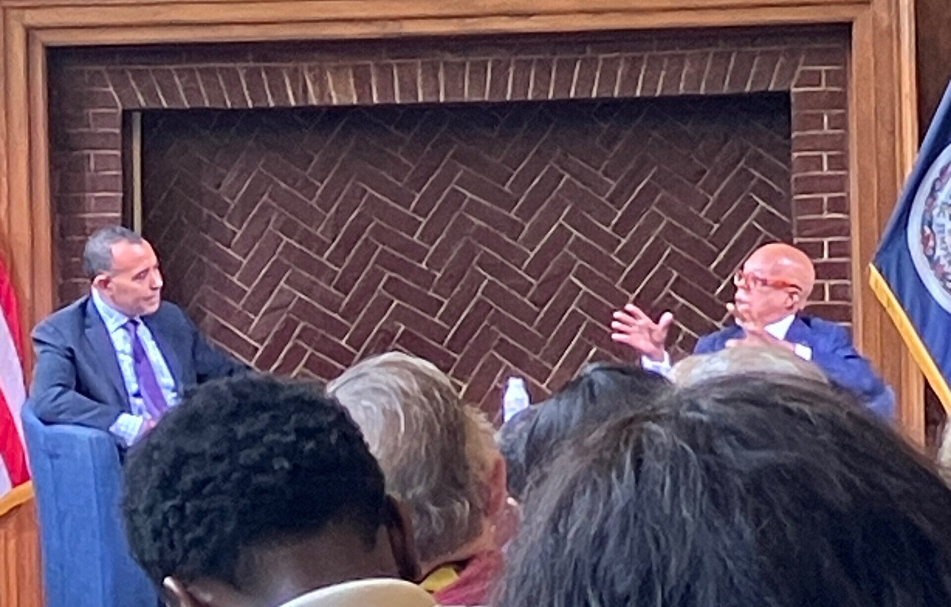 Two men sitting in front of a large empty fireplace, talking to each other in front of an audience.