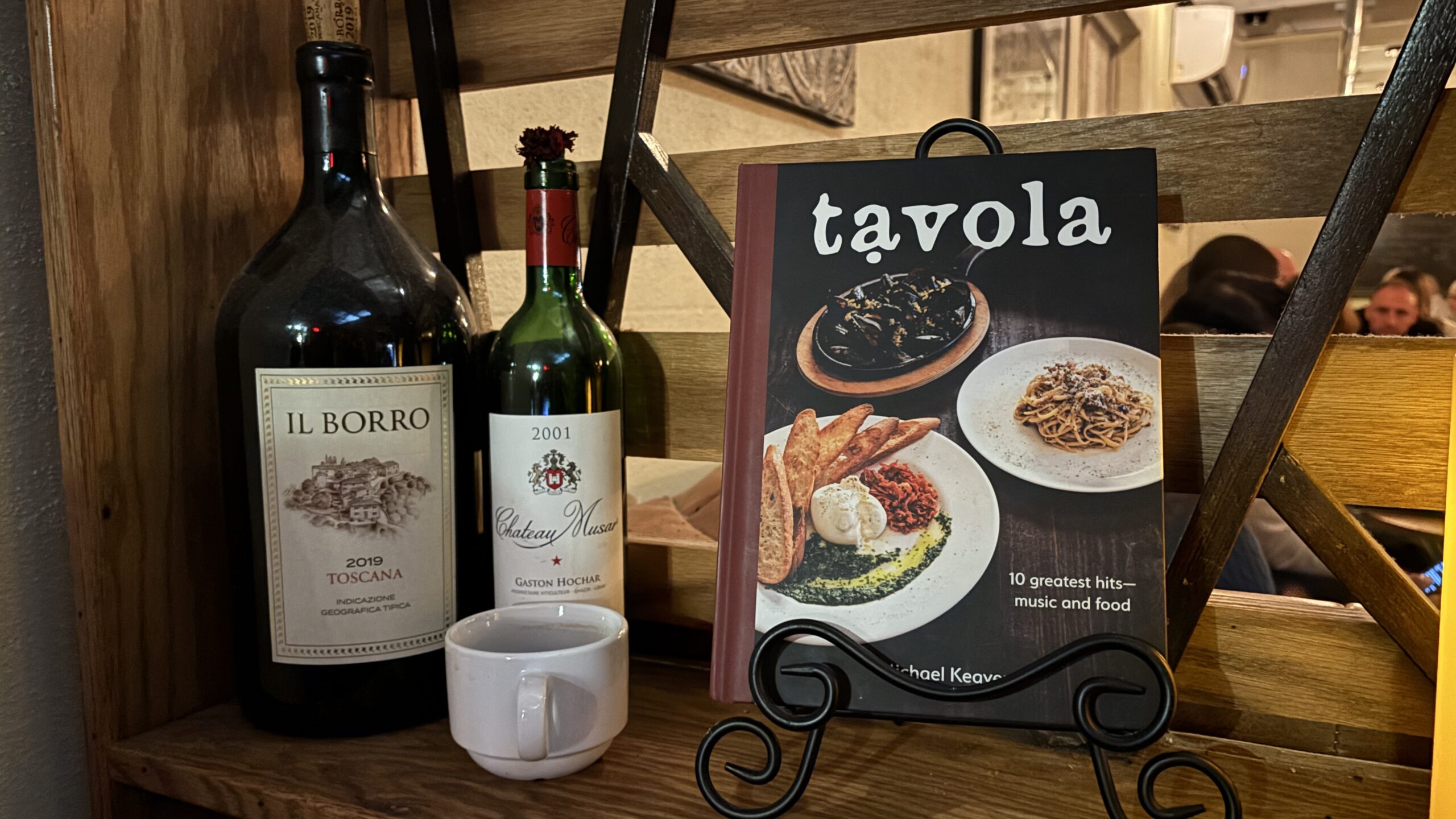 Two bottles of wine, an espresso mug, and a Tavola cookbook displayed behind the host stand at Tavola.