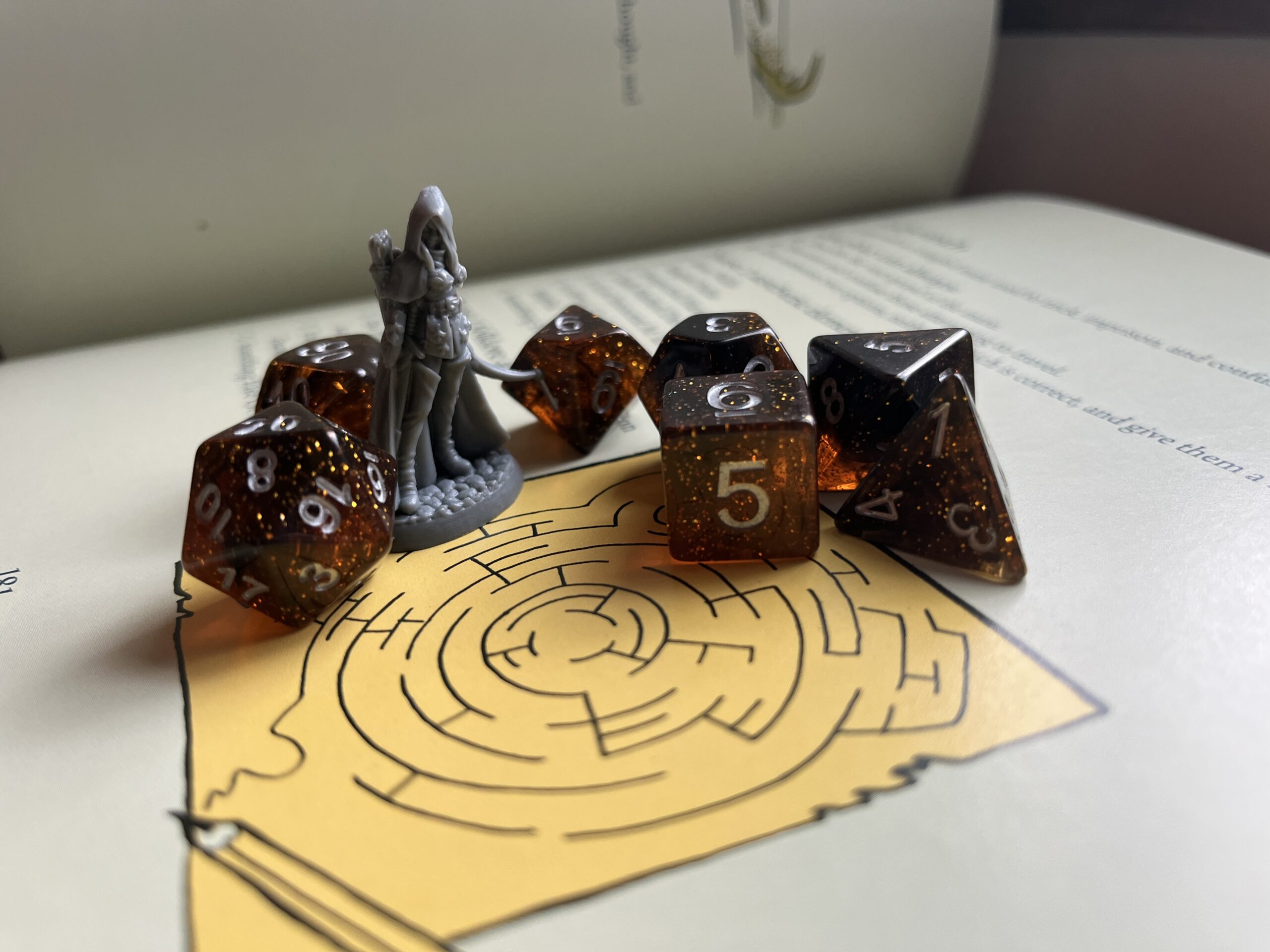 A set of seven tabletop role-play gaming dice ranging from four to 20 sides. They are sitting alongside a figurine on top of a maze on the page of an open book.