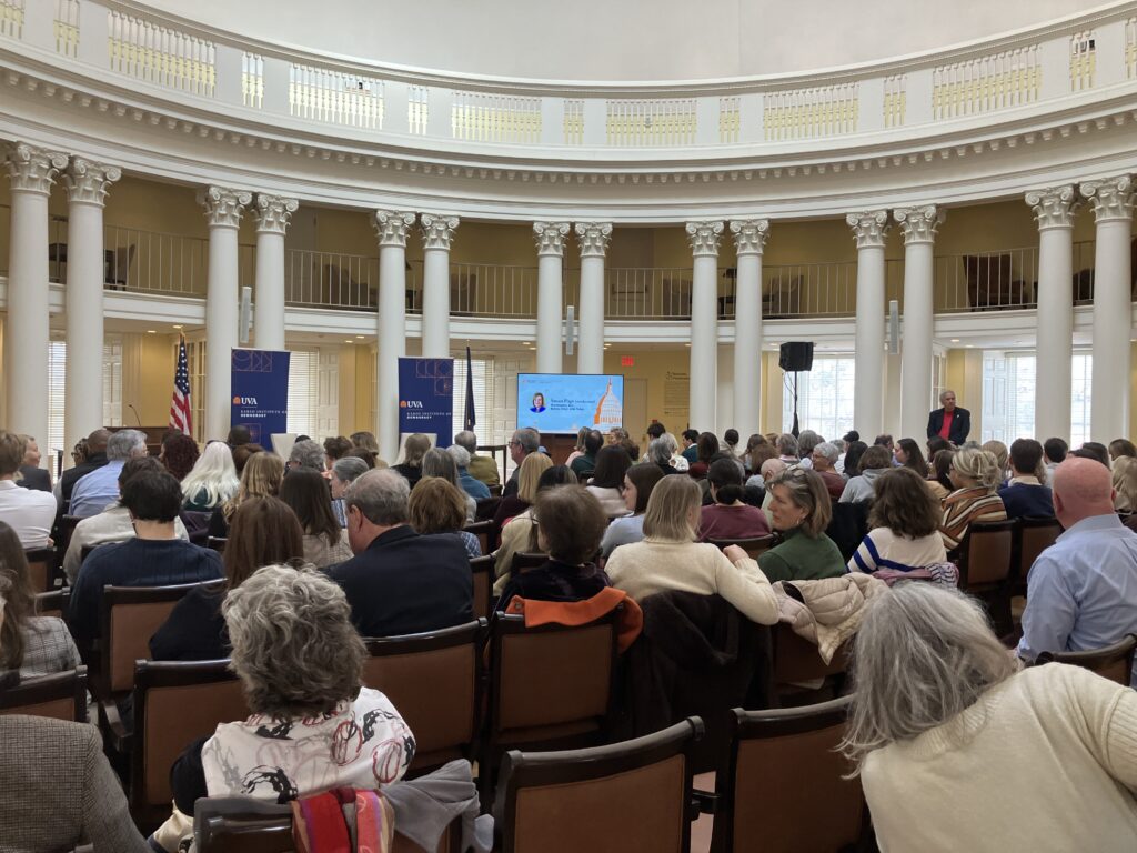 A large, circular room with a domed ceiling, balconies, and columns, with a full audience facing two empty seats in between blue banners and flags, with a television screen on the right. 