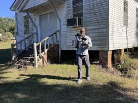 Ty Cooper standing in front of an old, white house holding up a high-tech camera.