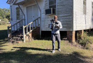 Ty Cooper standing in front of an old, white house holding up a high-tech camera.