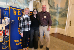 Morena Figueroa smiling with her host parents, Jim and Vicki Farmer, in front of a Rotary Club banner.