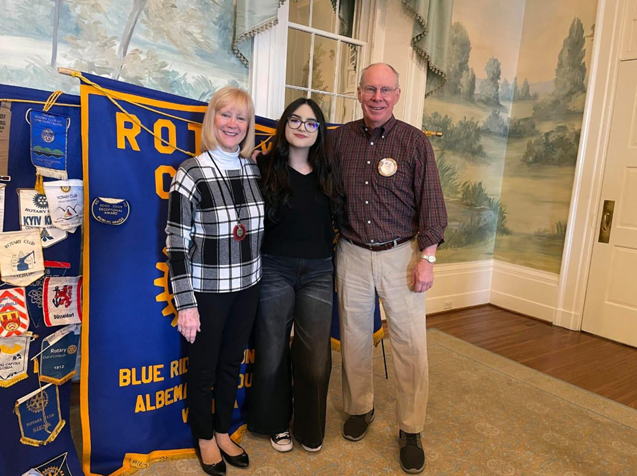 Morena Figueroa smiling with her host parents, Jim and Vicki Farmer, in front of a Rotary Club banner.