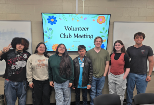 Seven volunteer club members stand smiling in front of a screen reading "Volunteer Club Meeting".