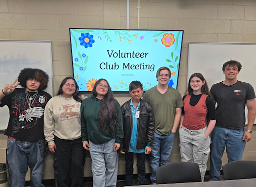 Seven volunteer club members stand smiling in front of a screen reading "Volunteer Club Meeting".